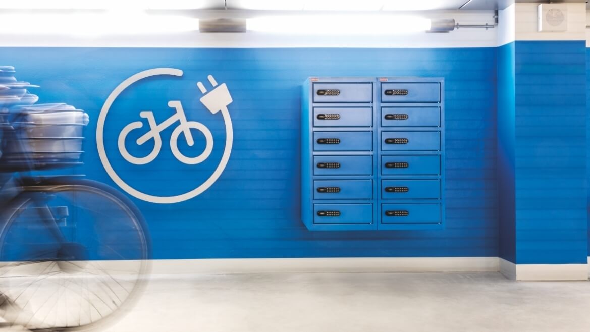 En gang med blå væg med ladeskabe til elcykler og et stort hvidt symbol for ladeskabe til cykler. En cykel kommer ind i billedet fra højre.